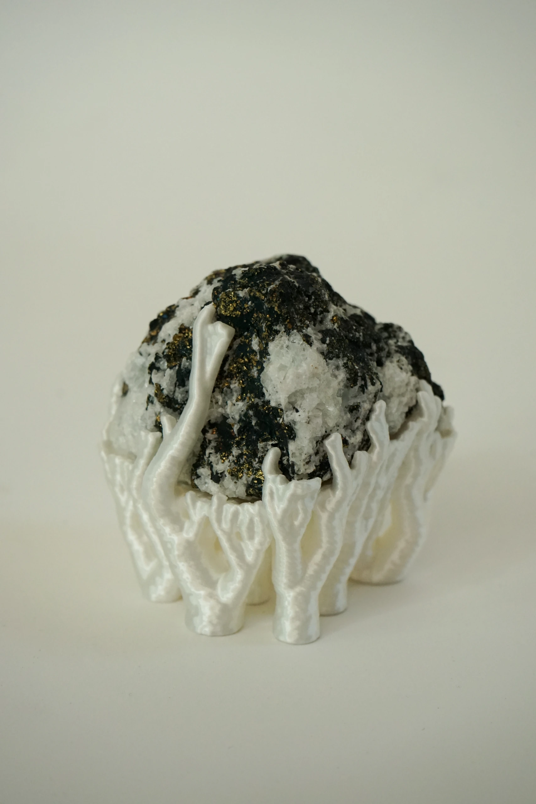 Mineral Cradle, sculpture by Sjors Driessen with a quartz-rich natural stone containing dark and metallic mineral inclusions, held by white branching hyphae-like structures.